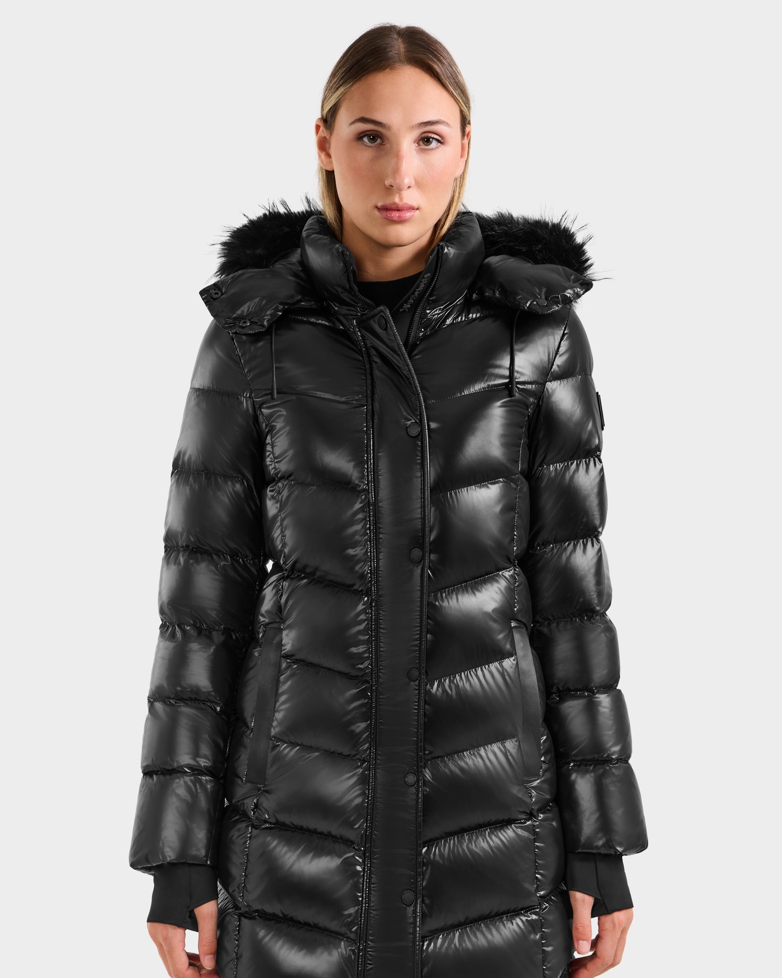 A woman models the Rudsak ELISA S - L125572 Black, a shiny black quilted puffer jacket with a faux fur-lined hood, standing against a plain light gray background.