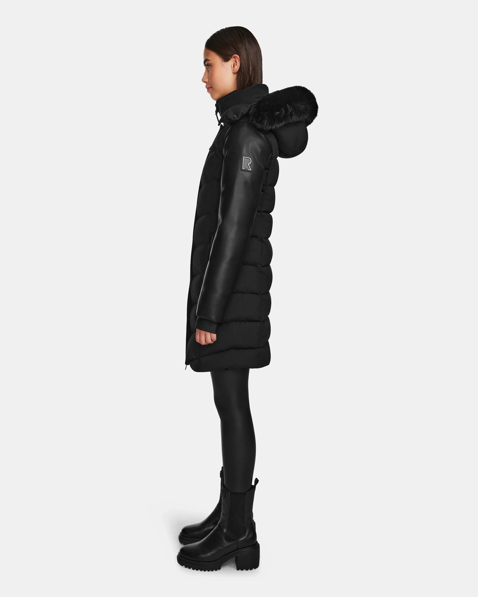 A person stands in profile wearing the Rudsak ELSA - L123535 Black womens quilted down puffer coat, paired with black leggings and boots, against a plain white background.