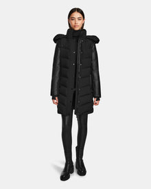 A woman faces forward in the Rudsak ELSA - L123535 Black, a sustainable long black women’s quilted down puffer with high collar and hood, styled with black leggings and ankle boots against a plain white background.
