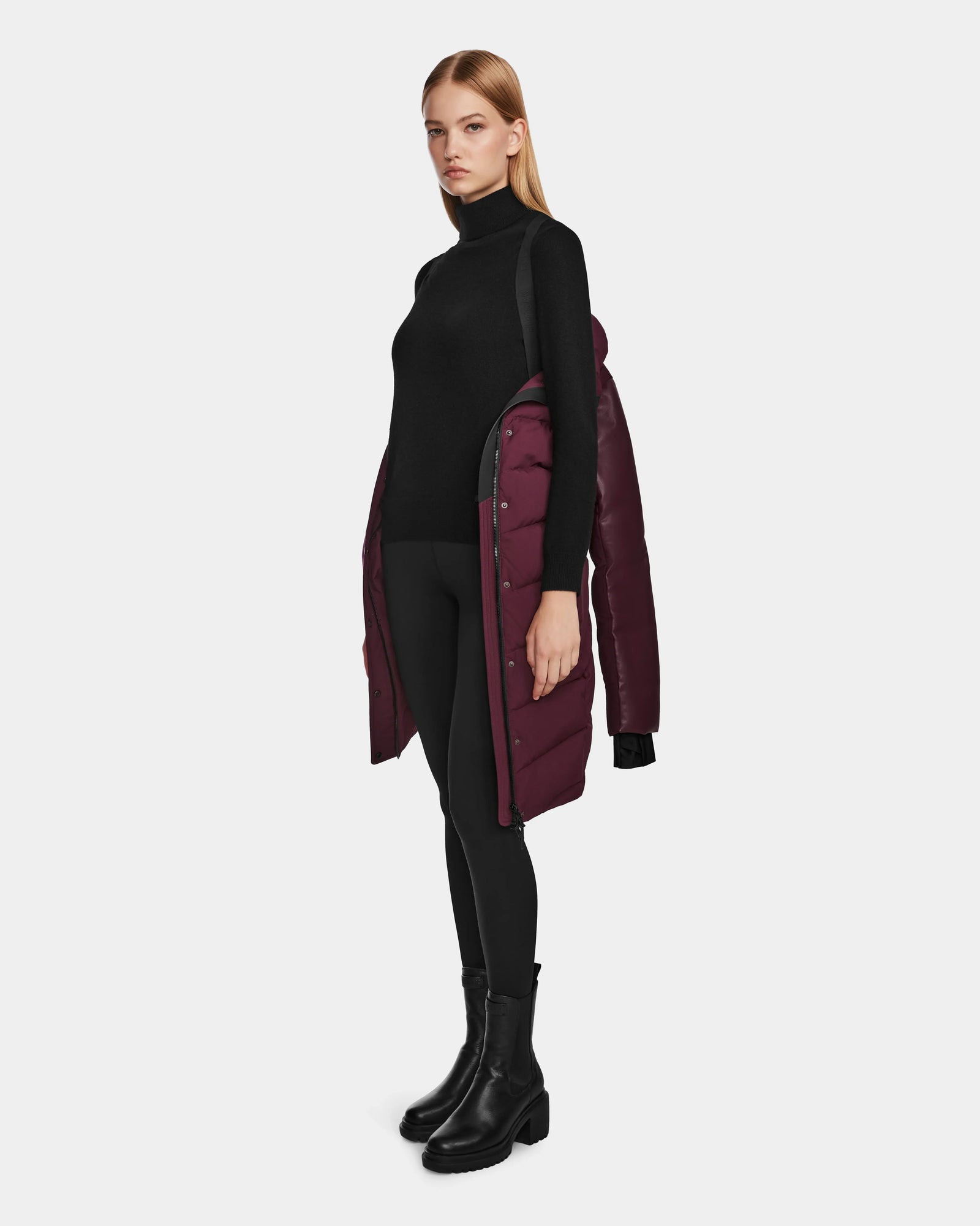 A woman with straight blonde hair stands against a plain light gray background, wearing a black turtleneck, leggings, and boots. She drapes the Rudsak ELSA - L123535 Wine women’s quilted down puffer over her shoulders.