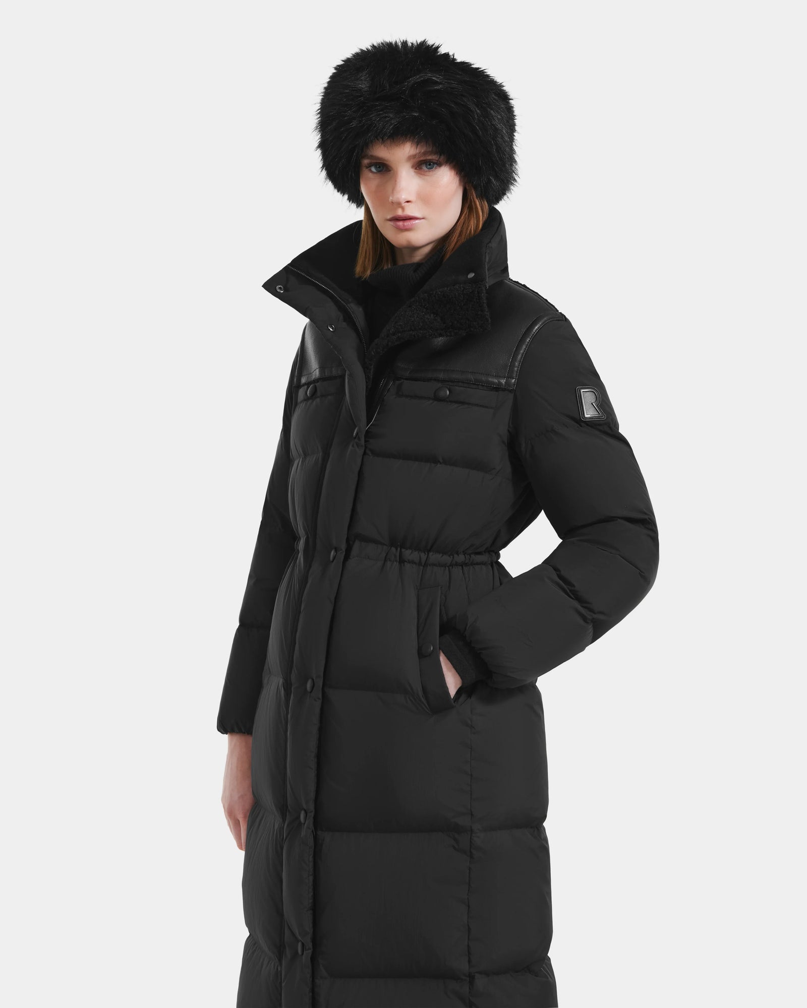 A woman stands against a plain light background wearing Rudsak’s EMBER M-L - L125543 Black women’s long down puffer coat with her hands in the pockets and a black fur hat.