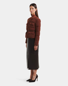 A woman stands sideways in a Rudsak FRANCESCA - 8125021 Rust fitted, down-quilted puffer jacket with a vegan leather belt, styled with a black midi skirt and high heels, against a plain white background.