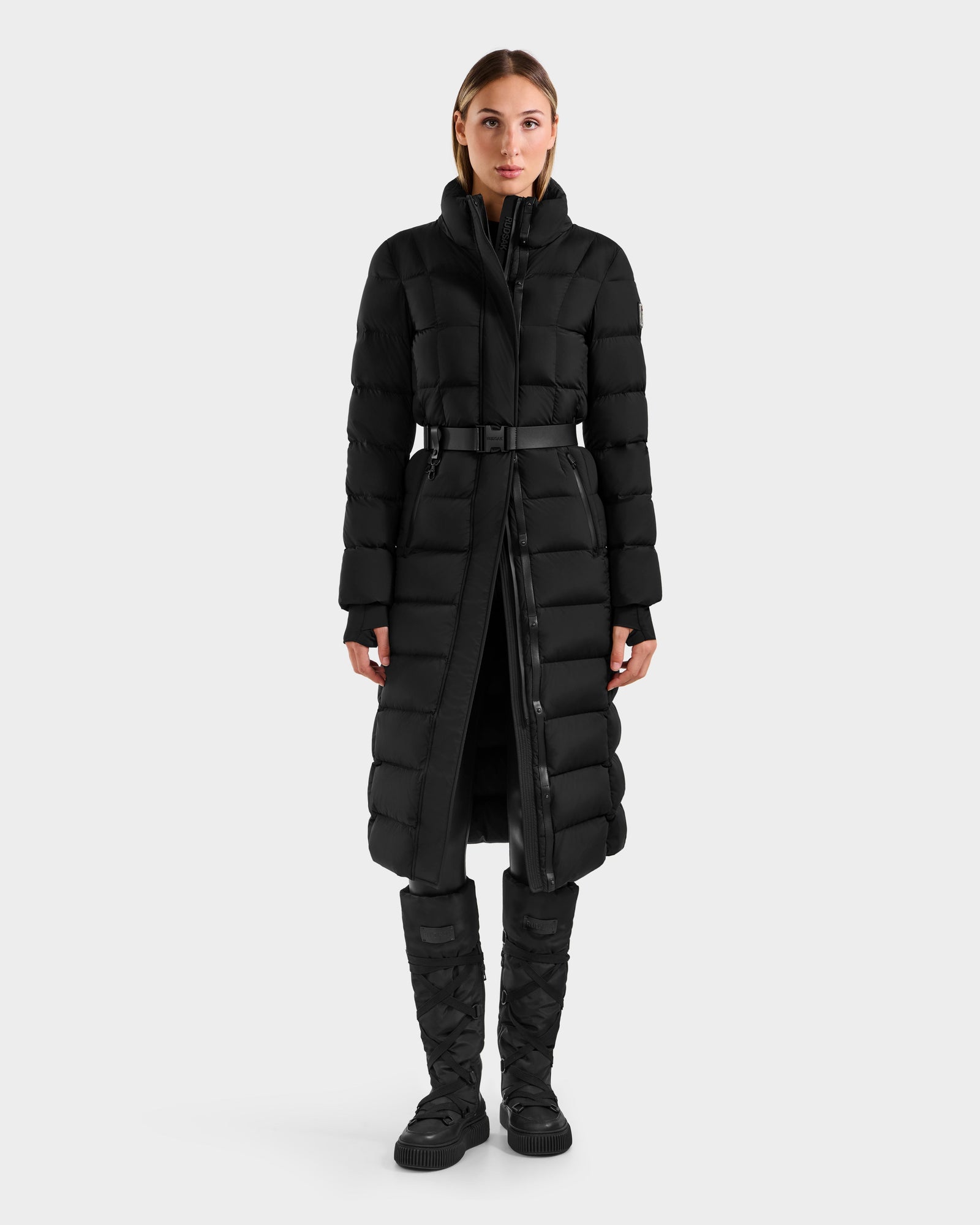A woman stands facing forward in the Rudsak FRIDA M-L - L125608 Black belted, quilted down puffer coat with a high collar and snap buttons, paired with black quilted boots, set against a plain white background.