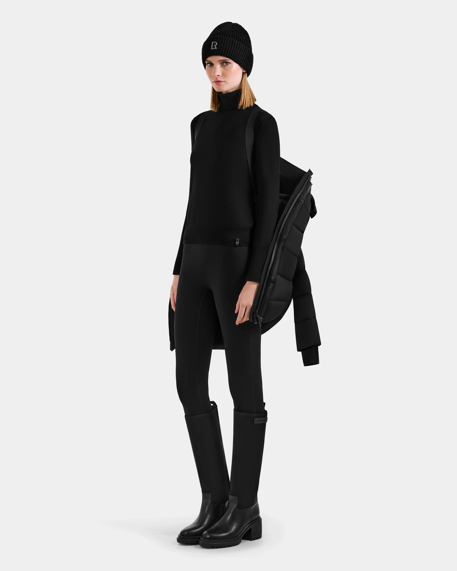 A woman in all-black attire stands confidently against a plain light background, featuring the Rudsak FRIDA M-M - L125607 Black quilted down puffer with detachable hood draped over her shoulder.