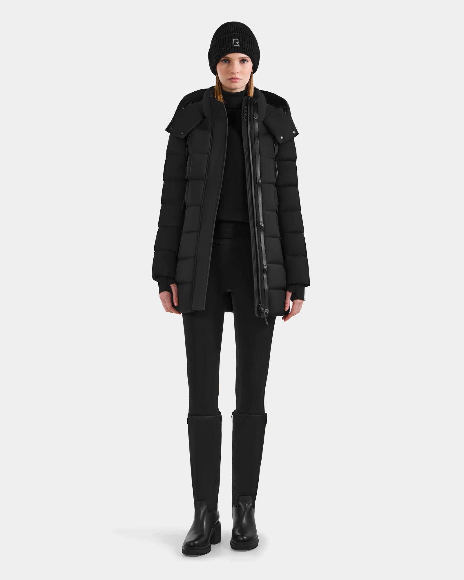 A person stands facing forward in the Rudsak FRIDA M-M - L125607 Black women’s quilted down puffer with detachable hood, paired with black pants and boots, against a plain light gray background—a true winter essential.
