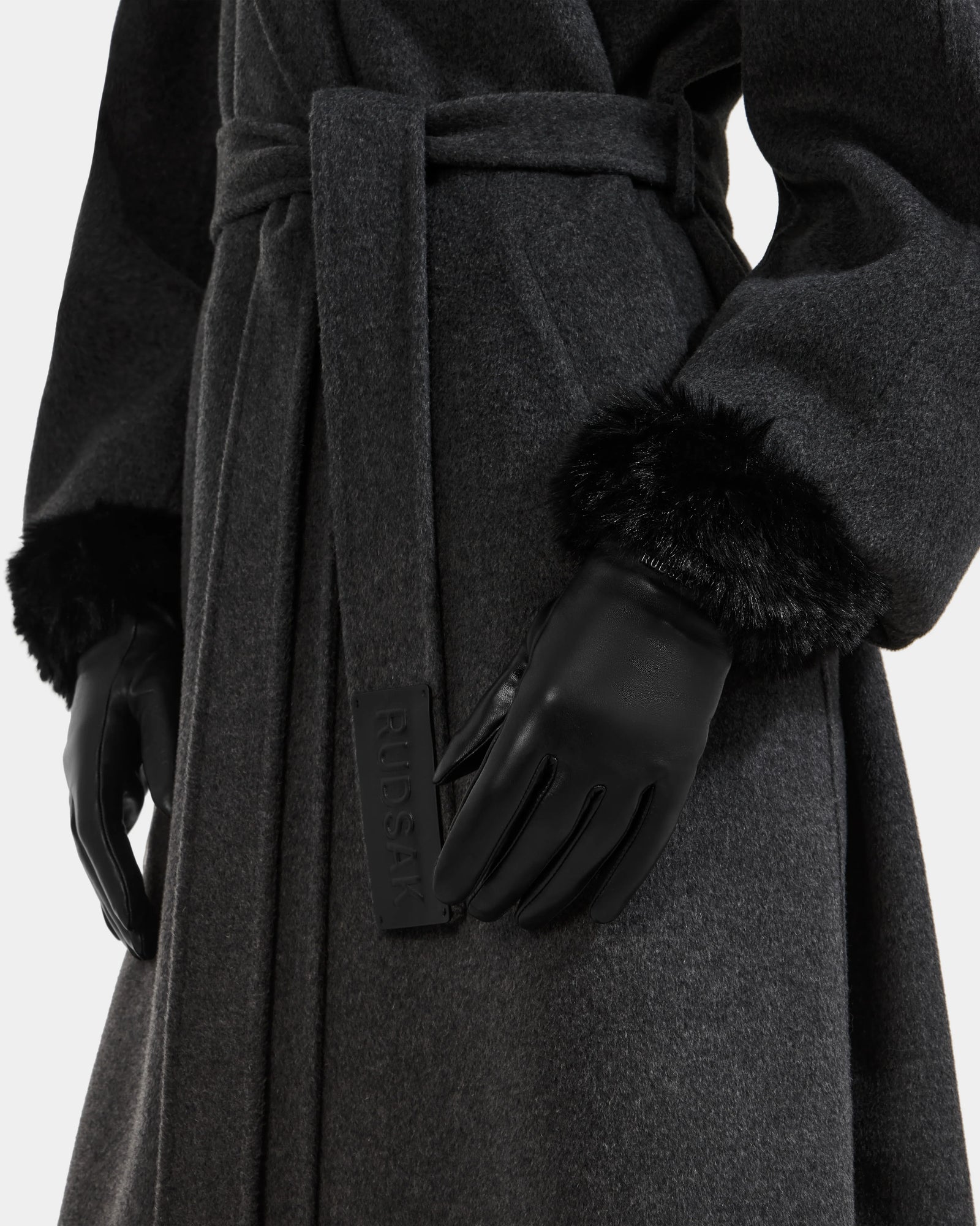 A close-up of a person wearing the Rudsak GERRY - L425550 Black belted coat and black faux-fur cuff gloves with a Rudsak tag. The women’s leather gloves add elegance against the plain white background.