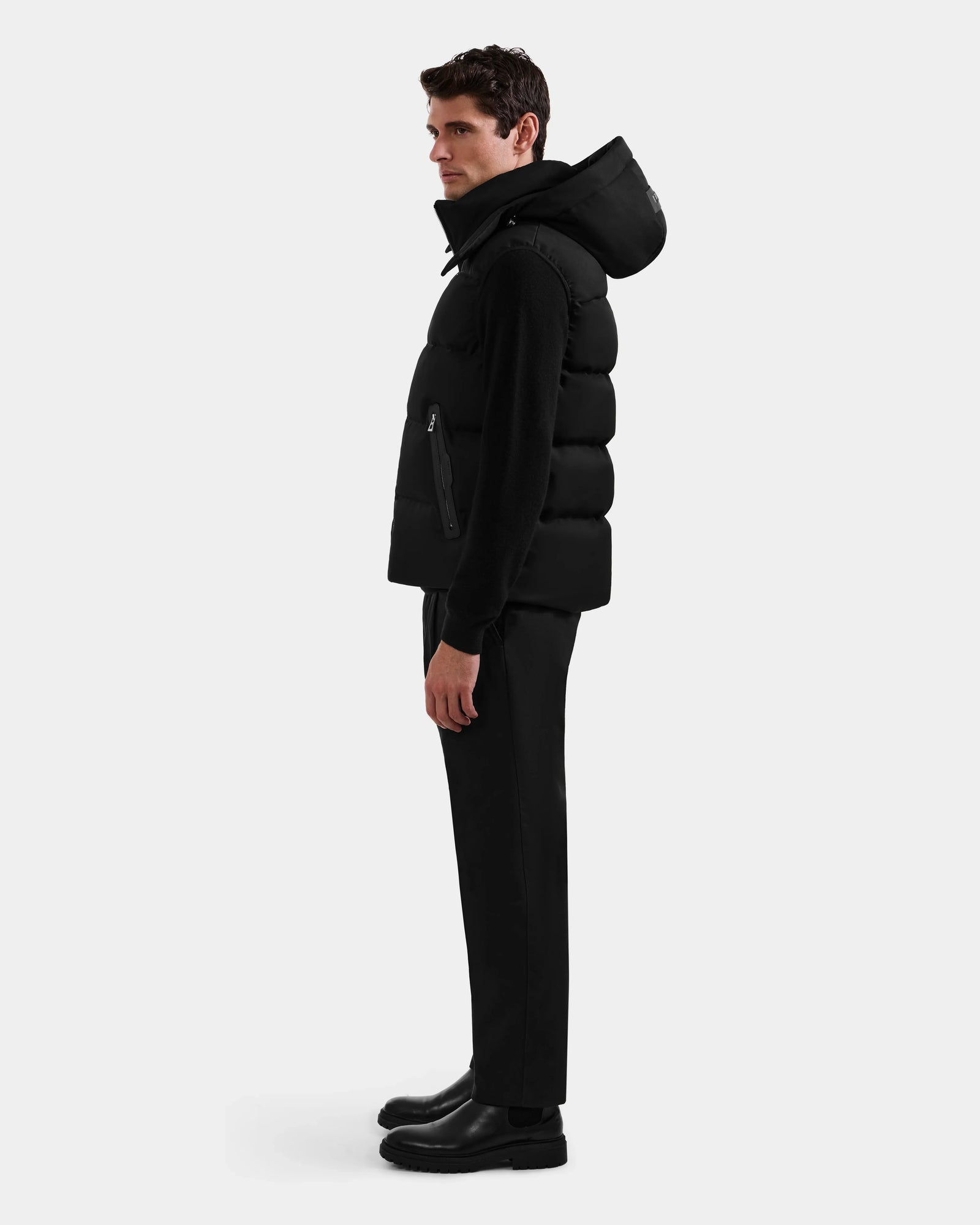 A man stands in profile against a plain background, wearing the Rudsak HARLOW - M125528 Black men's puffer vest with a removable hood, paired with black pants and boots.