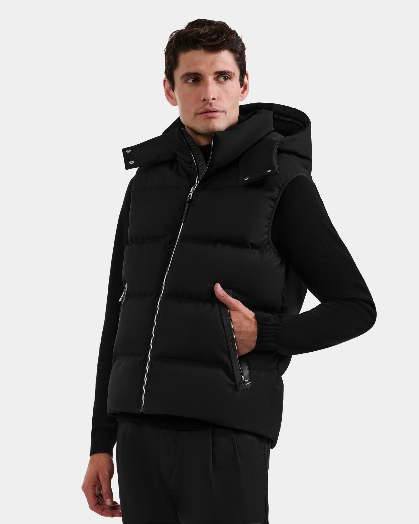 A man wears the Rudsak HARLOW - M125528 Black puffer vest with a removable hood over black attire, standing against a light background with one hand in his vest pocket, looking slightly to the side.