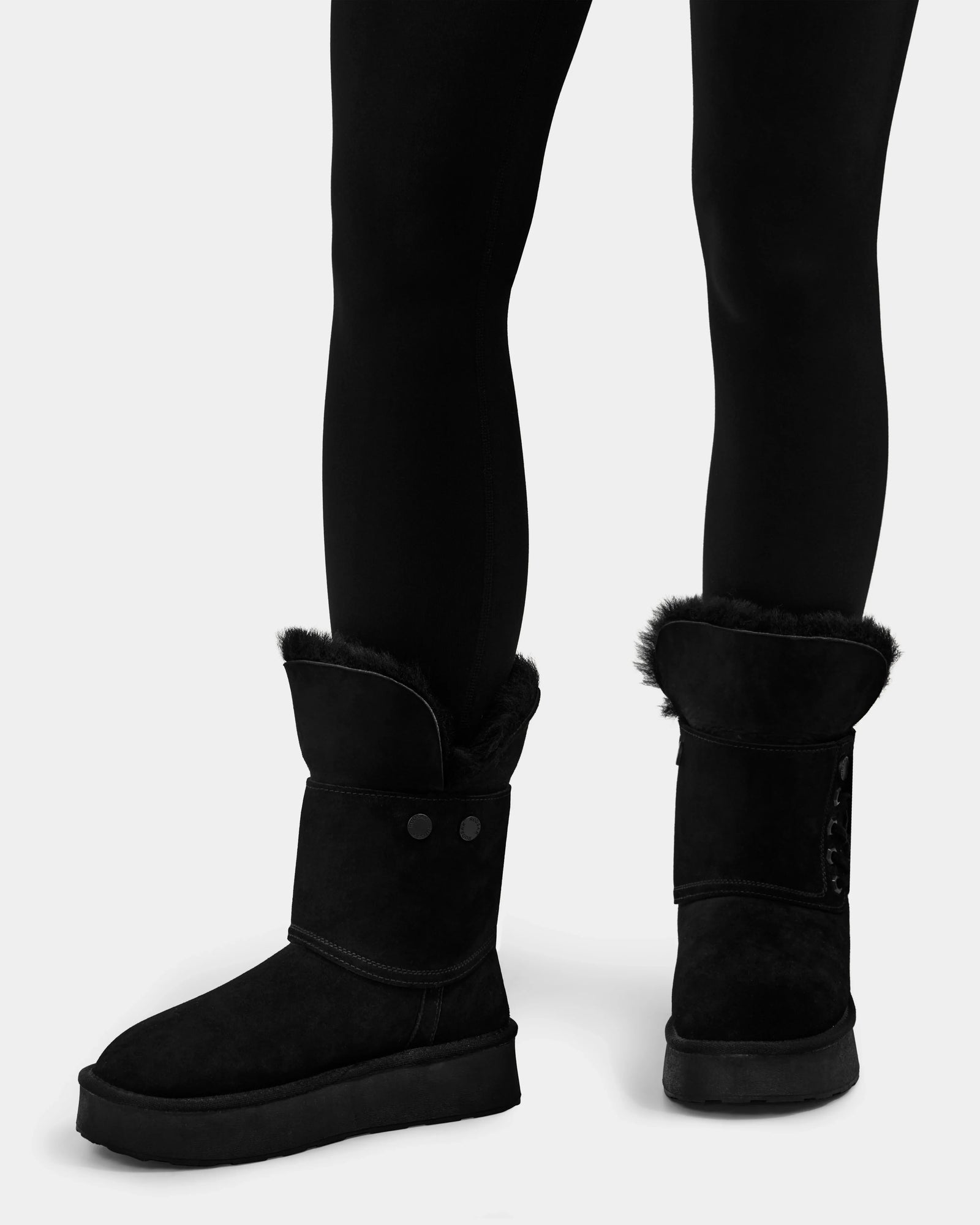 A person in black leggings and Rudsak INESA - L225026 Black women's suede platform boots with faux fur, wool lining, button details, and a lightweight sole stands against a plain white background.