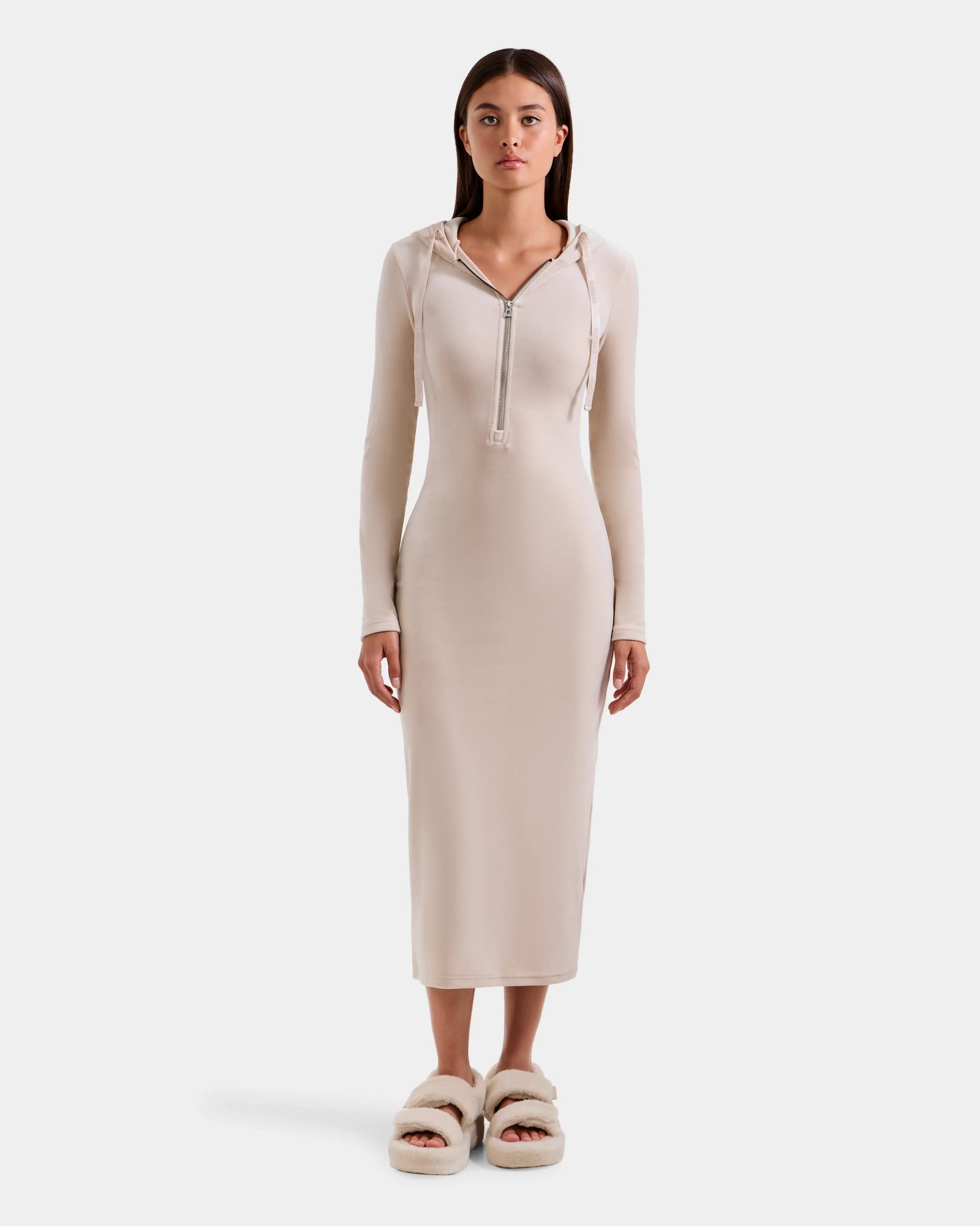 A woman faces forward in Rudsak’s ISABELLA - L525502 Cream: a chic, fitted velour hooded midi dress with long sleeves, front zipper, and matching fluffy slippers against a plain light background.