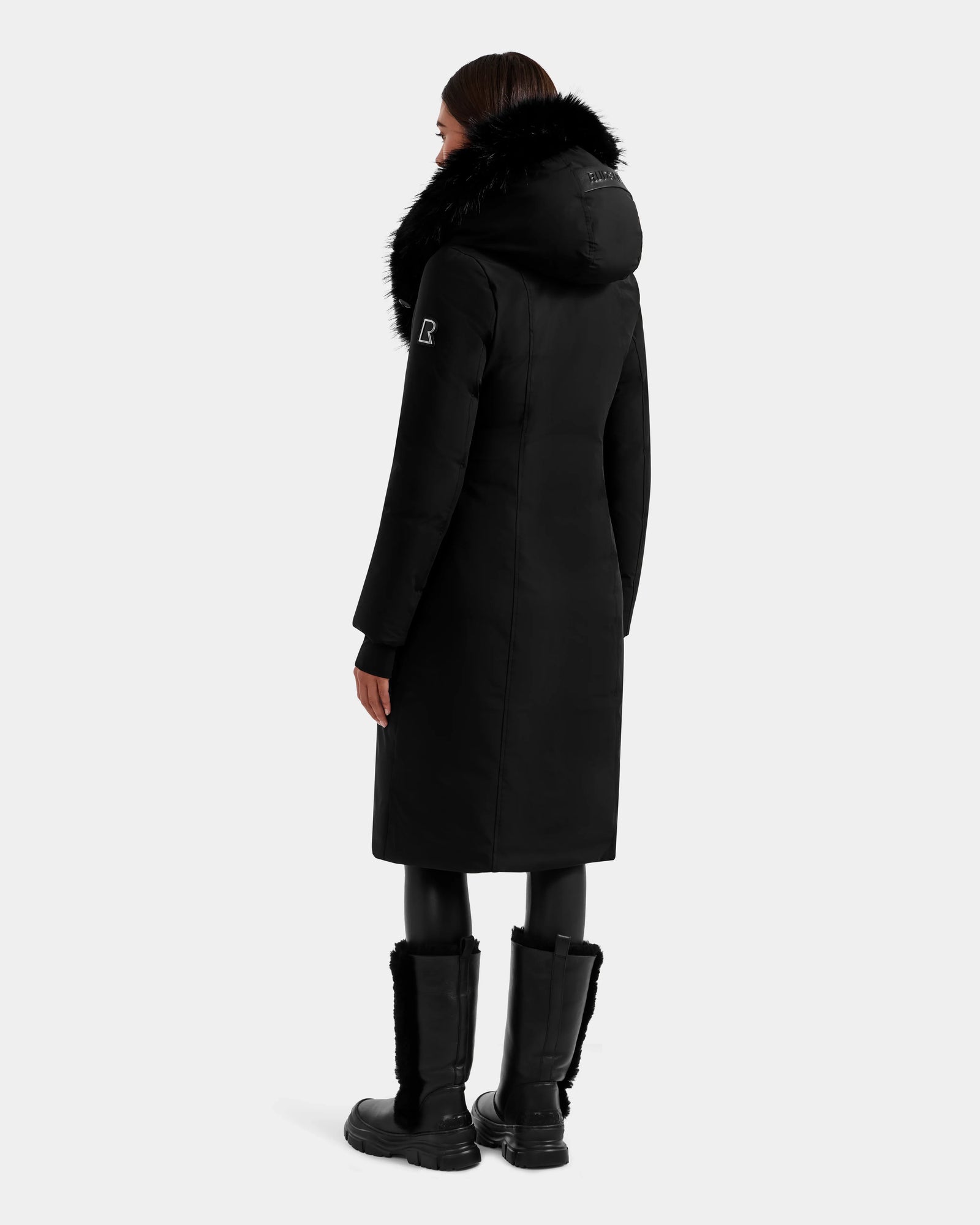 A person is standing and facing away from the camera against a plain light background, wearing the Rudsak JANIS - L125558 Black women’s down parka with a faux fur hood, black leggings, and black fur-trimmed boots.