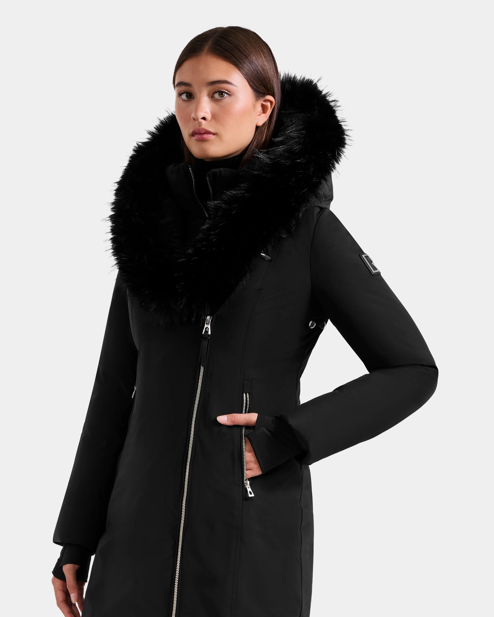 A woman wearing the Rudsak JANIS - L125558 Black long winter coat with a large, fluffy faux fur hood stands against a plain light background. This coat features a front zipper and zippered pockets for style and warmth.