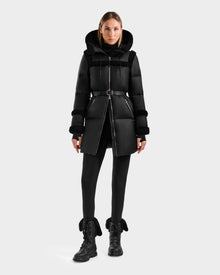 A woman faces forward in a Rudsak JULIANA L - L125535 Black/Black belted puffer coat with hood, black leggings, and black fur-trimmed lace-up boots, ideal for cold weather, set against a plain light gray background.