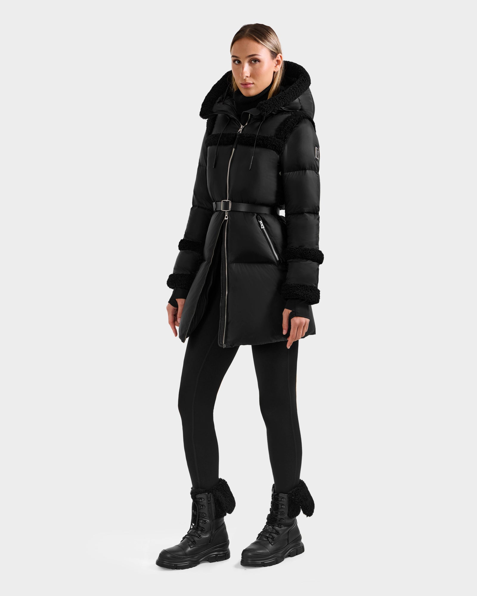 A woman with straight hair and a neutral expression stands against a plain background, wearing the Rudsak JULIANA L - L125535 Black/Black belted puffer coat with hood, paired with black leggings and lace-up boots. Hands by her sides.