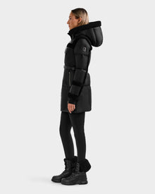 A woman in the Rudsak JULIANA L - L125535 Black/Black down puffer with a high collar, black leggings, and winter boots stands in profile against a plain light background, ready for cold-weather performance.