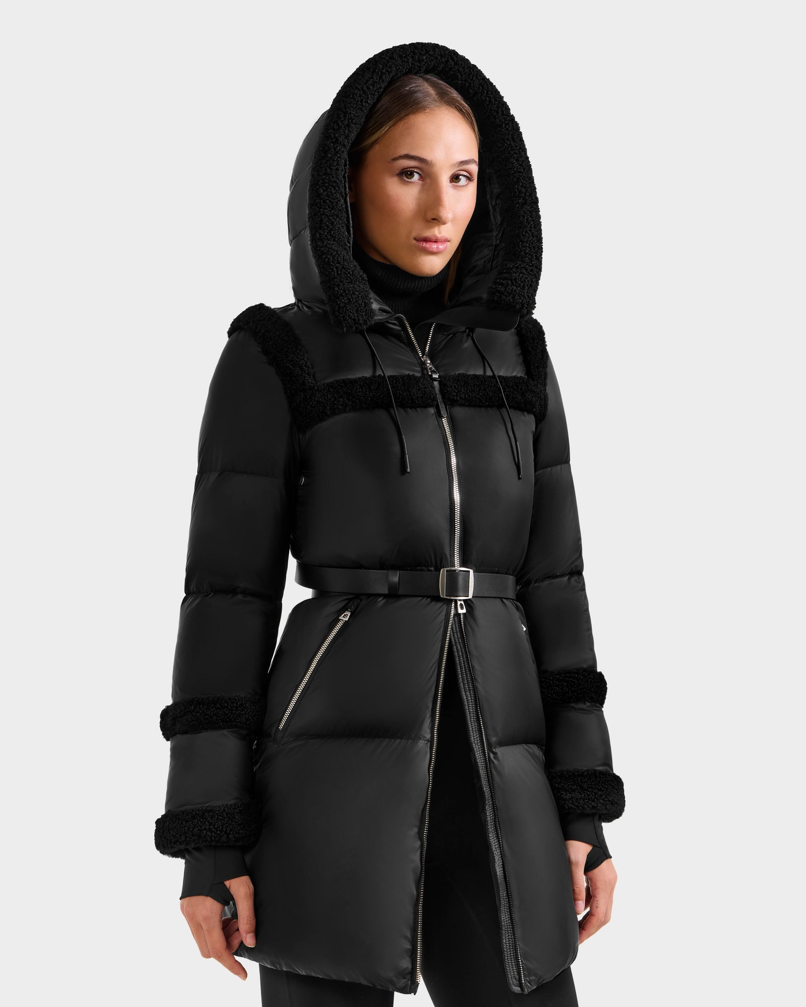 A woman wearing the Rudsak JULIANA L - L125535 Black/Black belted winter coat with a hood, faux fur trim, and RDS-certified down stands against a plain light gray background.