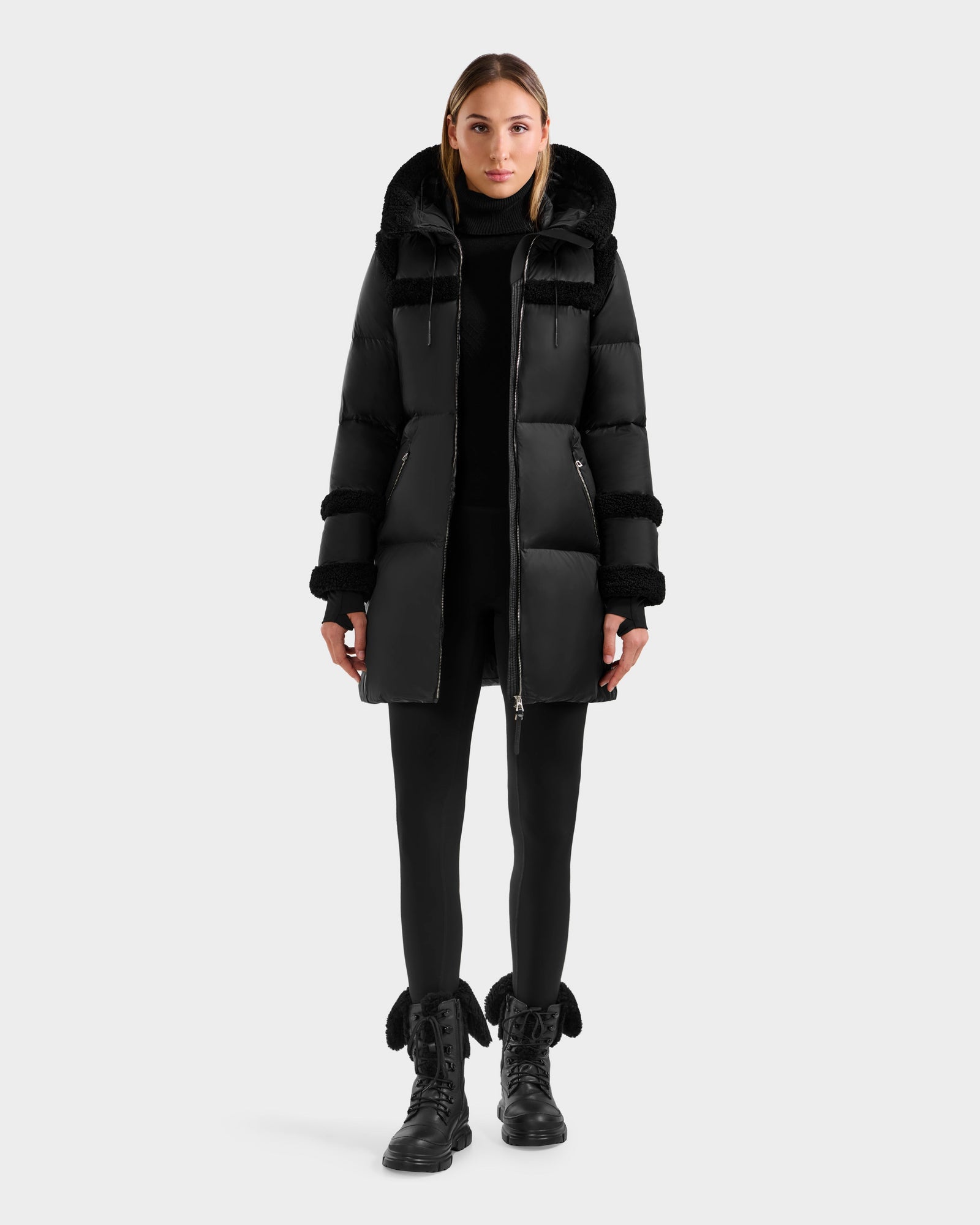 A woman faces forward wearing the Rudsak JULIANA L - L125535 Black/Black down puffer coat with fur trim, paired with black leggings and black lace-up boots, set against a plain light gray background.