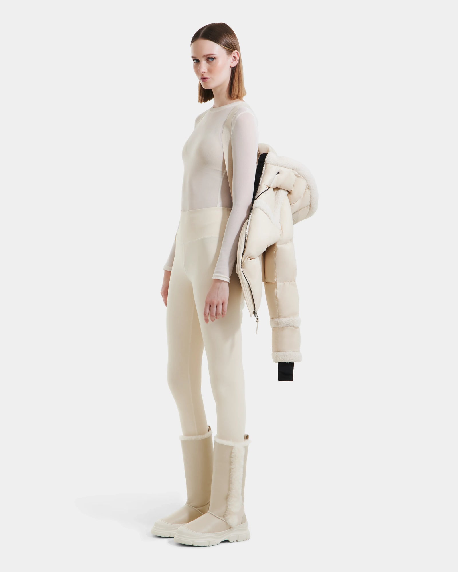 A woman stands against a plain white background, holding the Rudsak JULIANA - L124509 Vanilla women’s short down puffer over her shoulder—an ideal piece to elevate your winter wardrobe.