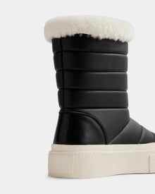 A close-up of Rudsak’s KATHIE - L225516 Black women’s mid boot, crafted from vegan leather with horizontal padding, a thick white sole, and fluffy white shearling trim, shown against a plain light background.