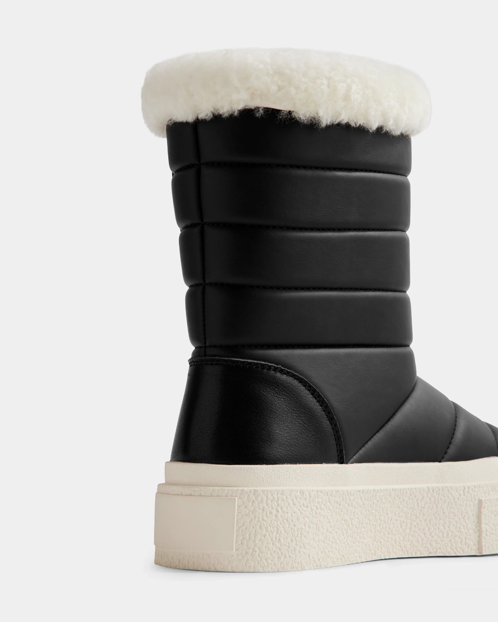 A close-up of Rudsak’s KATHIE - L225516 Black women’s mid boot, crafted from vegan leather with horizontal padding, a thick white sole, and fluffy white shearling trim, shown against a plain light background.