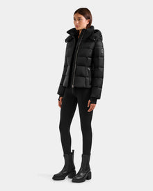 A woman with straight brown hair and a neutral expression stands against a plain background wearing the Rudsak KATIA - L125564 Black weather-resistant puffer jacket, paired with black leggings and ankle boots. Her hands rest by her sides.