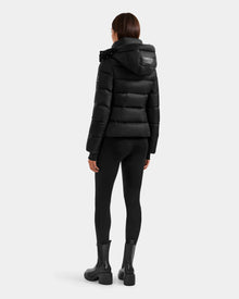 A person stands facing away, wearing the Rudsak KATIA - L125564 Black weather-resistant puffer jacket with a hood, paired with black leggings and ankle boots against a plain white background.
