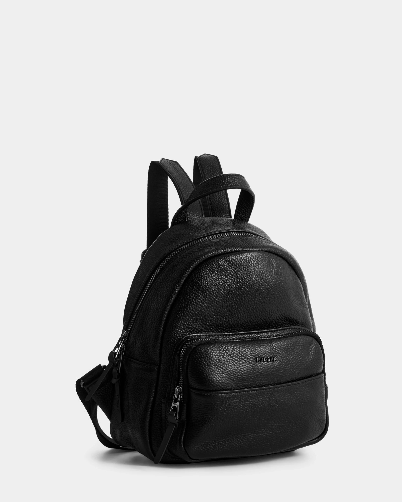 The Rudsak KATTY - L325014 Black is a women's mini backpack in black pebbled leather, featuring a front zip pocket, adjustable shoulder straps, and silver zippers, shown against a plain light gray background.