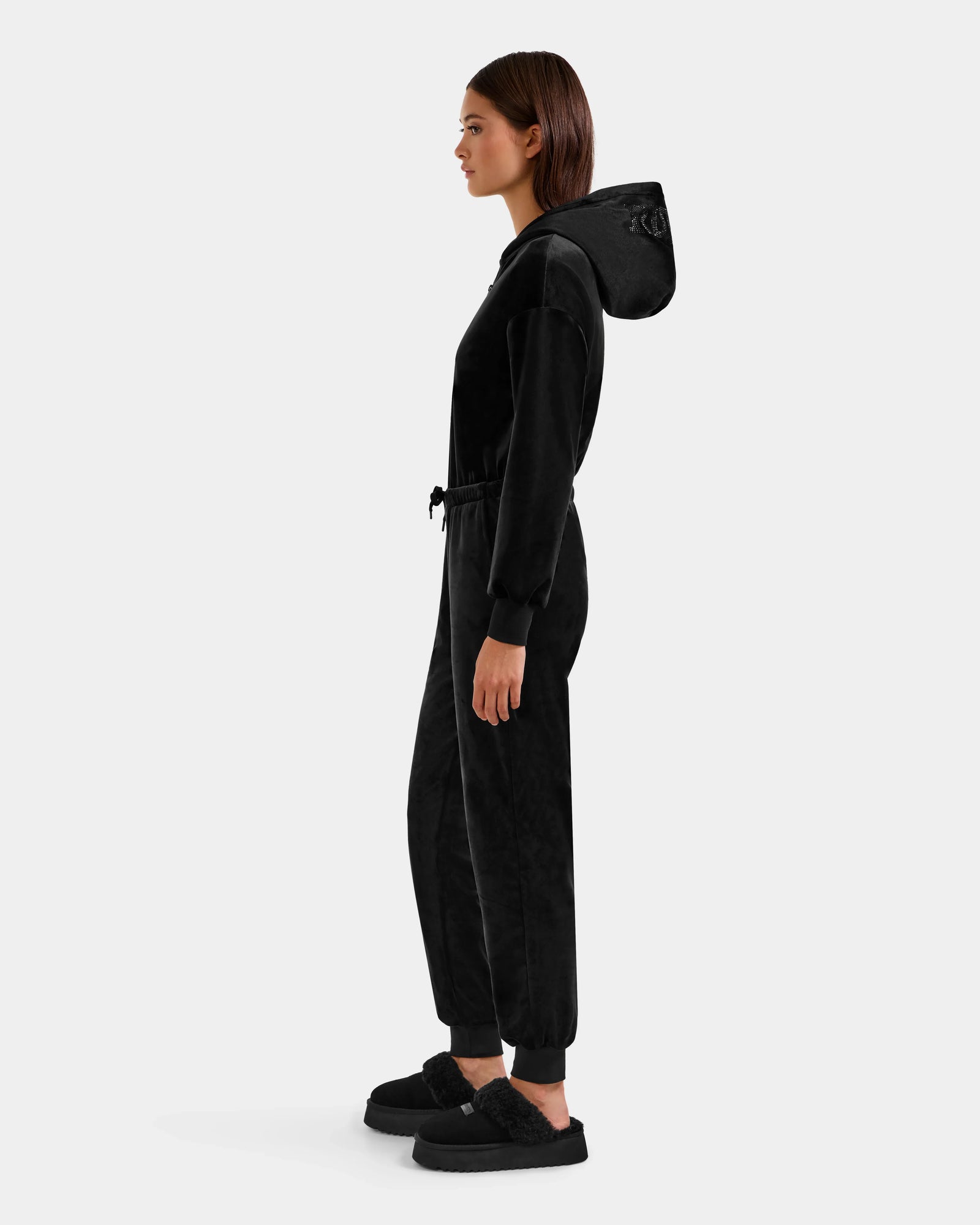 A woman stands in profile against a light gray background wearing the Rudsak KETZY - L525503 Black velour hooded jumper with a drawstring waist and black slippers, crafted from stretch velour fabric.