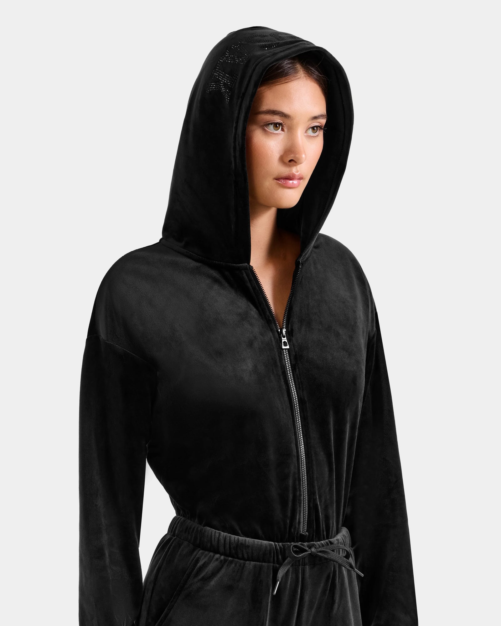 A woman wears the Rudsak KETZY - L525503 Black velour hooded jumper with matching drawstring pants, both featuring a crystal-sequin logo, as she stands against a plain light background.