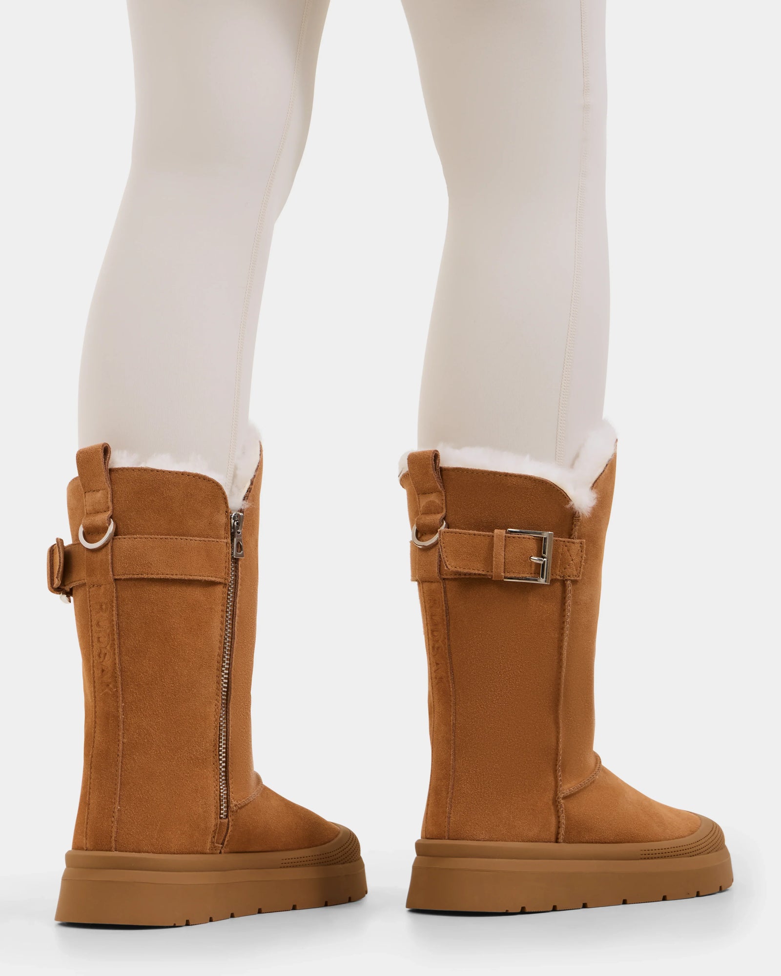 A person wears cream leggings and Rudsak’s KLAIR - L225027 Tan women's mid boots—tan suede with buckle, zipper details, and a white wool lining peeking out—standing against a white background.