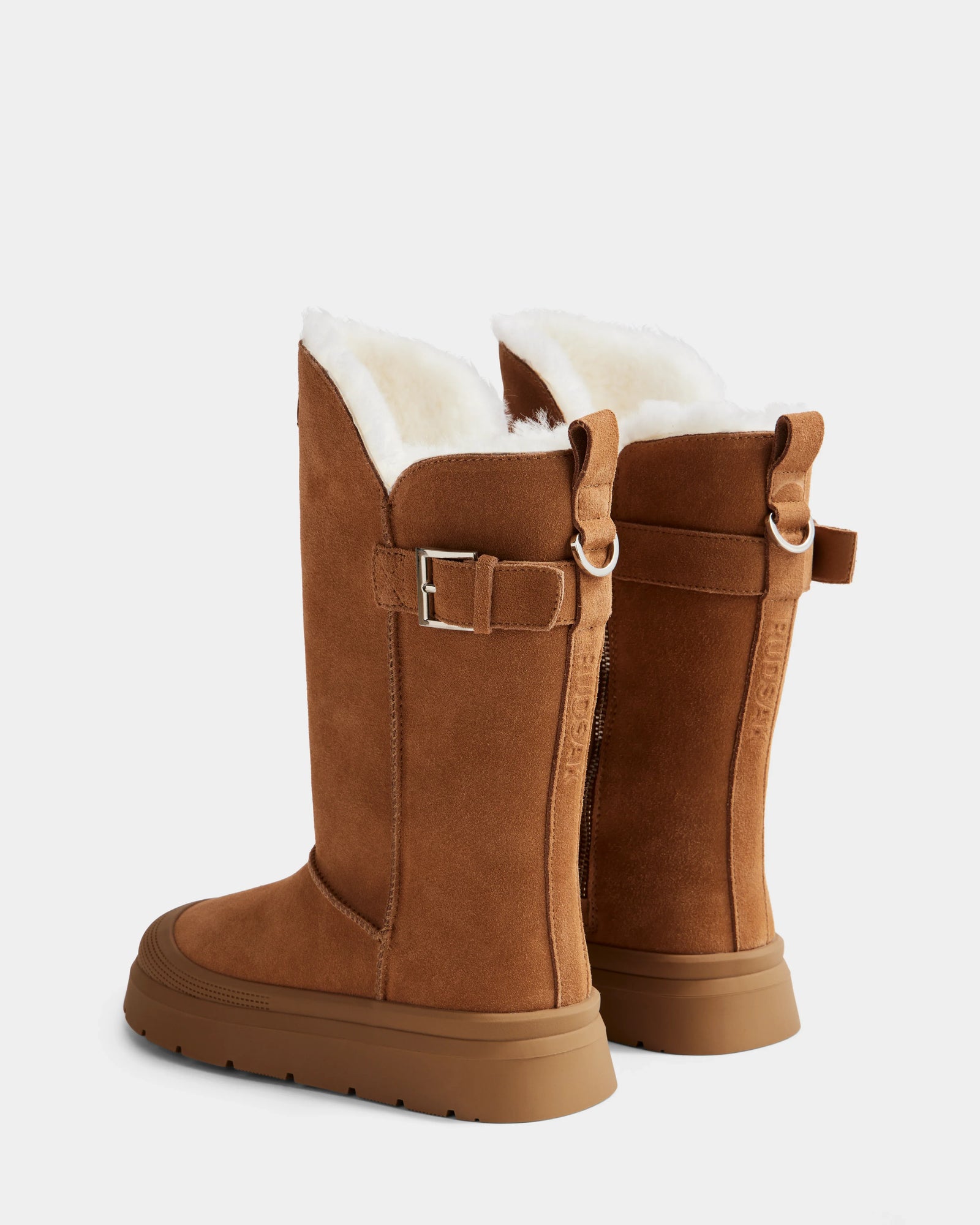 The Rudsak KLAIR - L225027 Tan features tall brown suede winter boots with white wool lining, side buckle detail, and flat rubber soles, shown from the back at an angle on a plain light background.