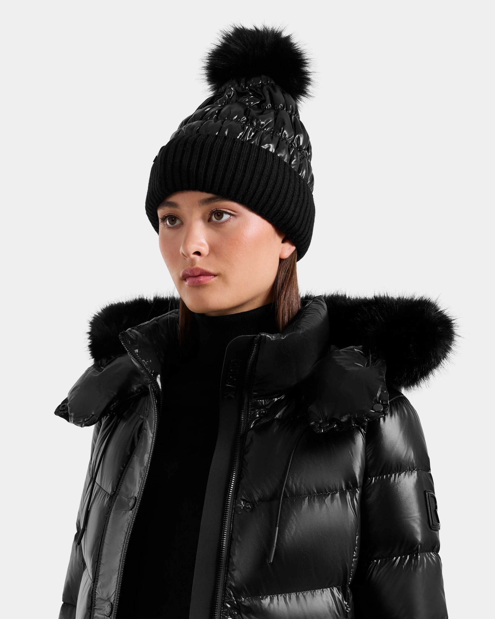 A woman wearing the Rudsak LANYA - L423514 Metallic Black puffer jacket and a women's pom pom hat, looking slightly to the side against a plain light background.