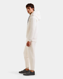 A man stands sideways against a plain light background, wearing the Rudsak LEROY - U525509 Vanilla M premium unisex hoodie, matching white pants, and black sneakers.