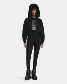 A person wears Rudsak's LIDDIE - L525505 Black: an all-black après-ski outfit featuring a furry trapper hat, SKI COUTURE women’s sweatshirt, fitted leggings, and puffy snow boots, standing against a white background.