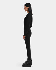 A woman with straight dark hair stands in profile, wearing Rudsak’s LIDDIE - L525505 Black fitted outfit with glossy black boots and a cropped crewneck sweater, capturing après-ski style against a plain white background.
