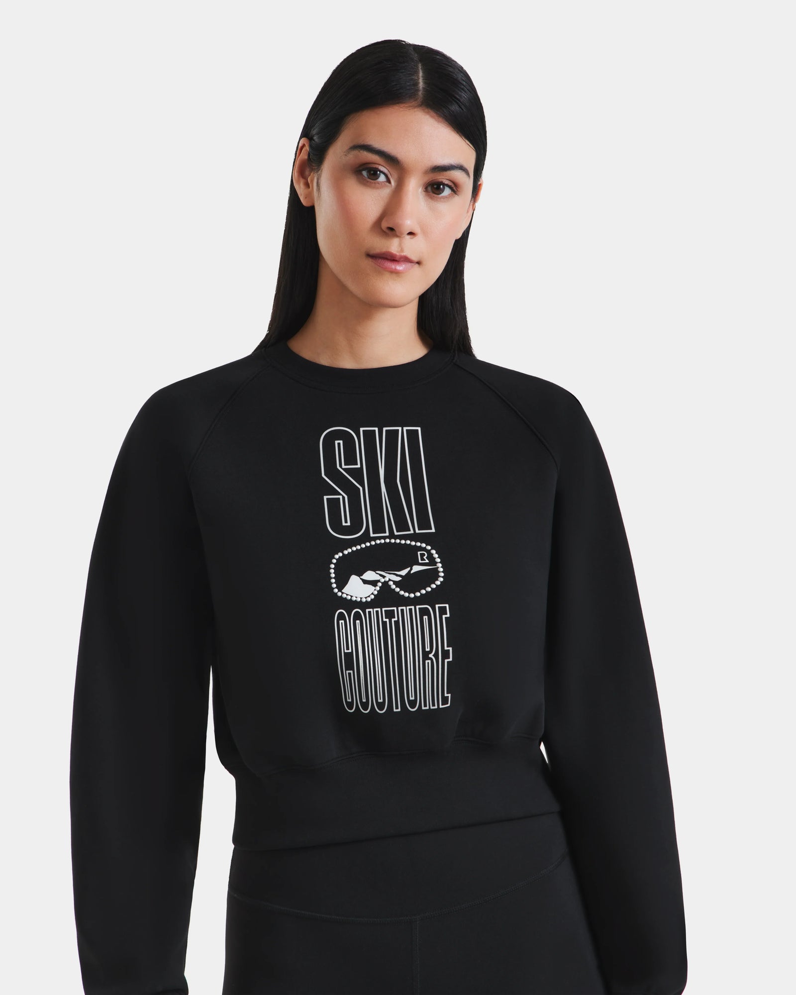 A woman with long dark hair wears the Rudsak LIDDIE - L525505 black cropped crewneck sweatshirt, featuring "SKI COUTURE" in white text and a goggles graphic, standing against a plain white background.