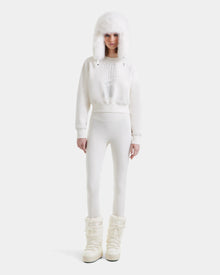 A person stands facing forward in the Rudsak LIDDIE - L525505 Vanilla: an all-white winter look with a furry hat, cropped crewneck sweater, fitted pants, and puffy high snow boots, set against a plain white background.