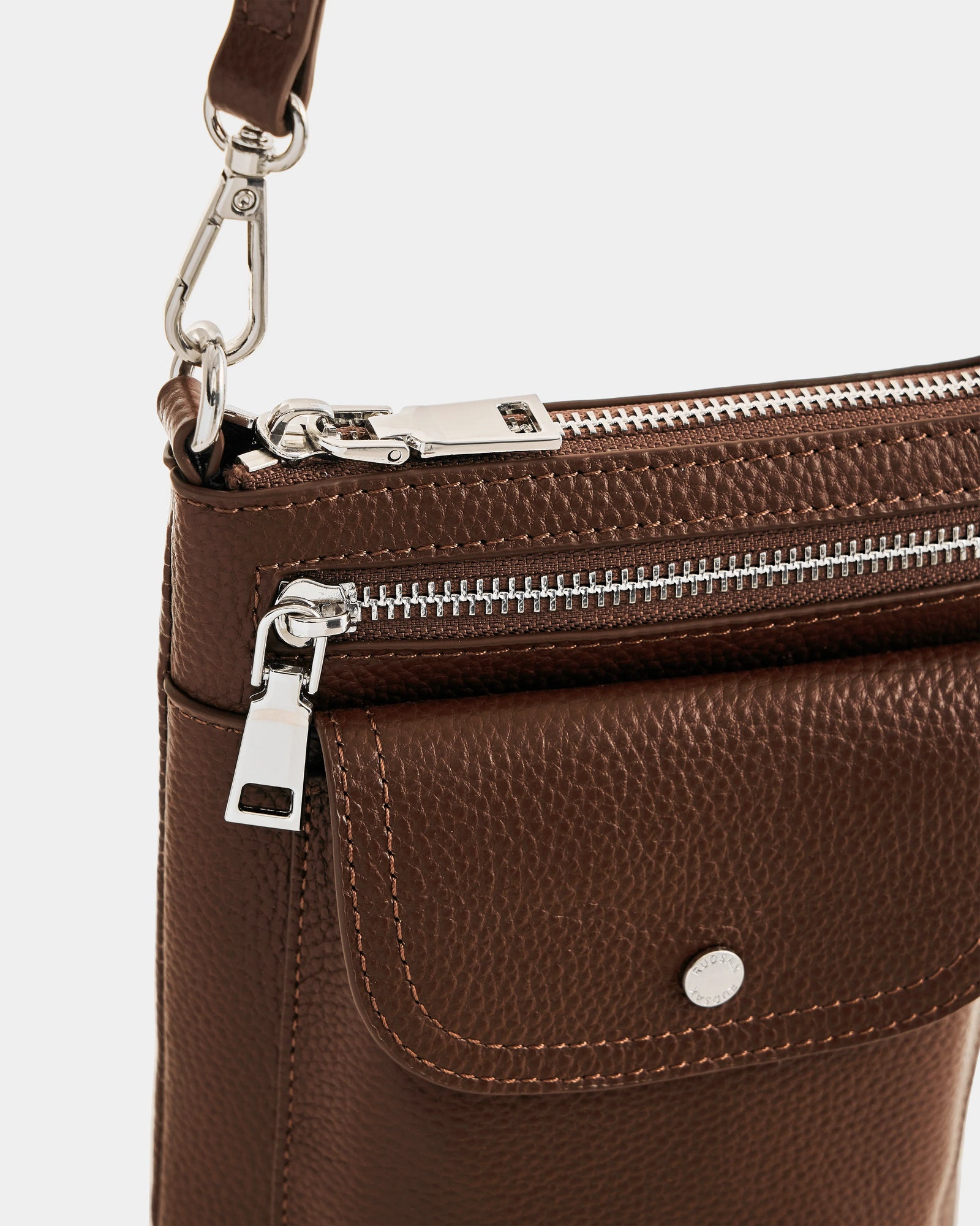 Close-up of the Rudsak LILA - L325513 Mocha women’s crossbody bag, featuring silver hardware, a front snap pocket, adjustable strap clip, and textured pebbled leather.
