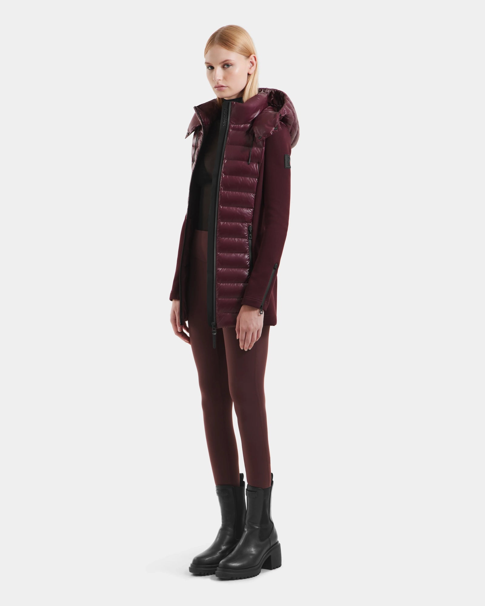 A woman with straight blonde hair models the Rudsak MARRY S - L125521 Wine glossy hooded puffer coat, styled with maroon pants, a black top, and heeled boots against a plain white background.