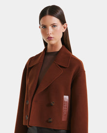 A woman with straight brown hair wears the Rudsak MARTINEZ - 8125580 Rust women’s short coat with wide lapels and a vegan leather RUDI SÈK patch over a sheer brown top, standing against a plain white background.
