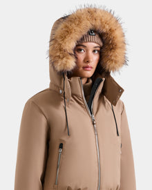 Wearing the MAUDE - L124522 Sand women’s down bomber by Rudsak, featuring premium down insulation and a fur-lined hood, paired with a brown knit beanie. The zipped-up jacket’s hood frames her face against a plain light background.