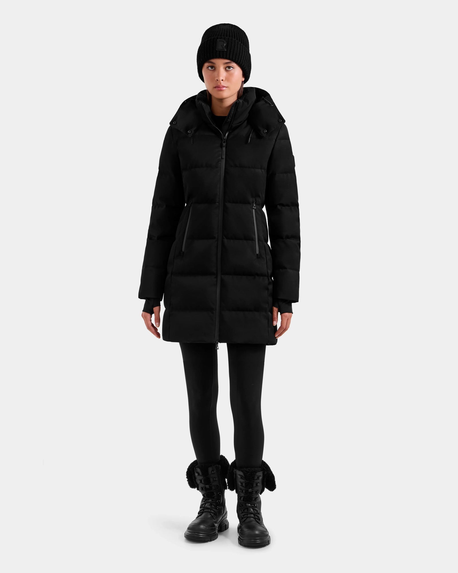 A person faces forward wearing the Rudsak MIRIAM MW - L125604 Black women's down puffer coat with RDS-certified down, paired with a black beanie, leggings, and lace-up boots against a plain light background.