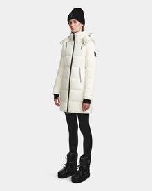 A person stands against a plain background, wearing the Rudsak MIRIAM - L123544 Iceland womens long white down puffer winter jacket with a high collar, zip pockets, sleeve logo, black leggings, black boots, and a black knit beanie.