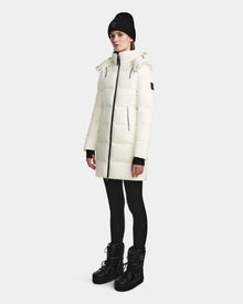 A person stands against a plain background, wearing the Rudsak MIRIAM - L123544 Iceland womens long white down puffer winter jacket with a high collar, zip pockets, sleeve logo, black leggings, black boots, and a black knit beanie.