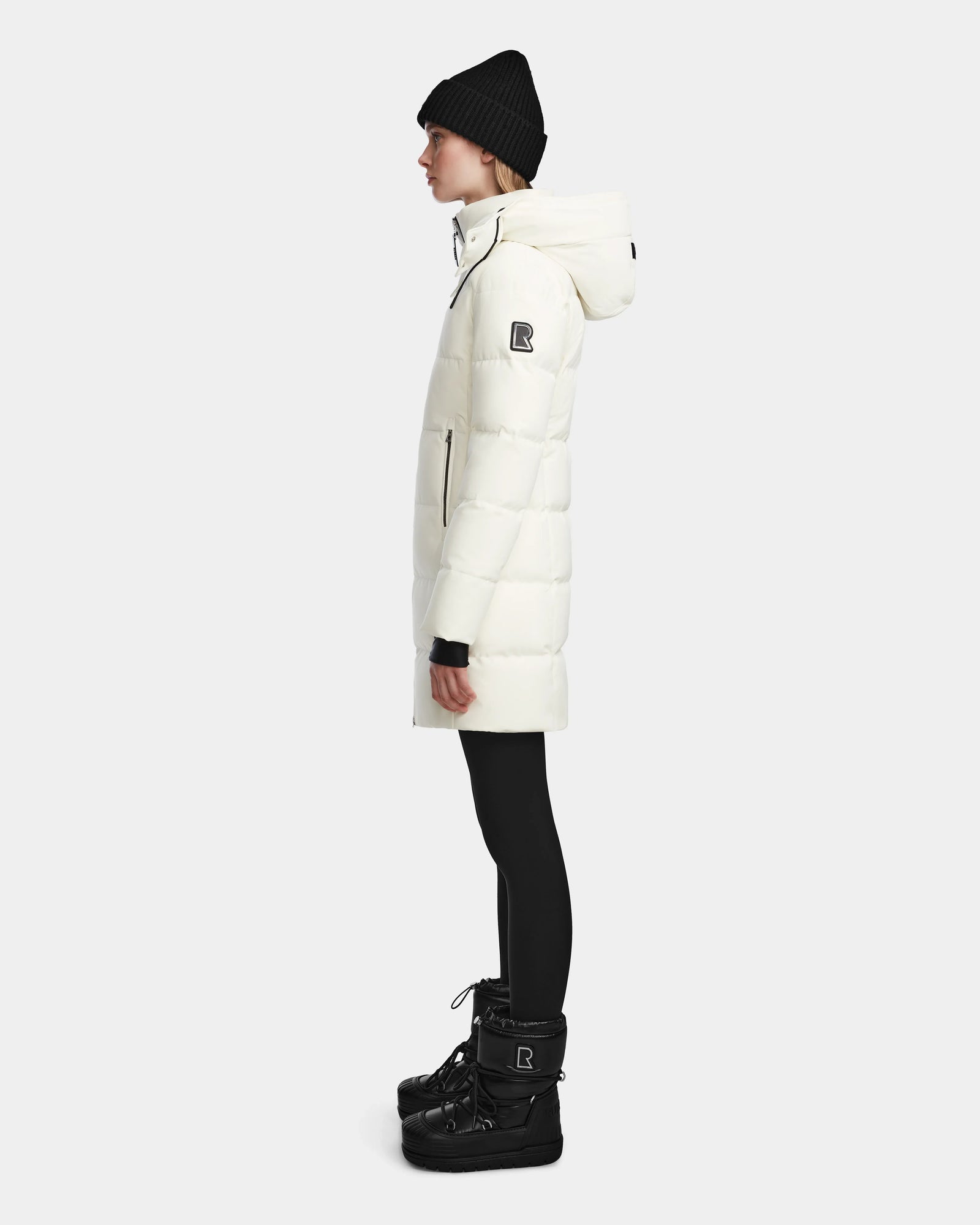 A woman stands in profile against a plain light background wearing the Rudsak MIRIAM - L123544 Iceland womens down puffer with a hood, paired with black leggings, winter boots, and a knit beanie—a stylish and practical cold-weather look.