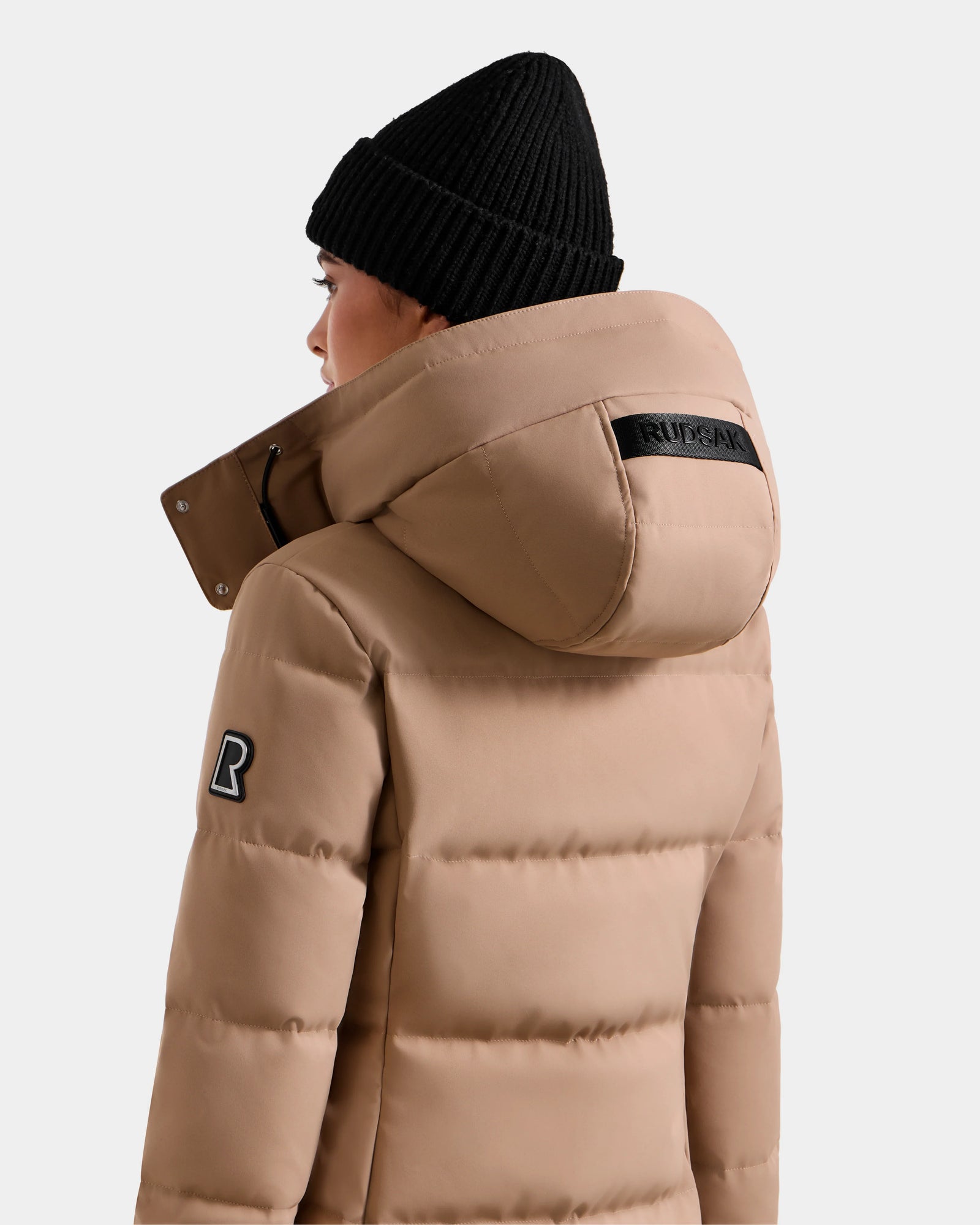A person is shown from behind wearing the Rudsak MIRIAM - L123544 Sand weather-resistant puffer jacket with a large hood, black beanie, a black text patch on the hood, and a logo patch on the upper sleeve.