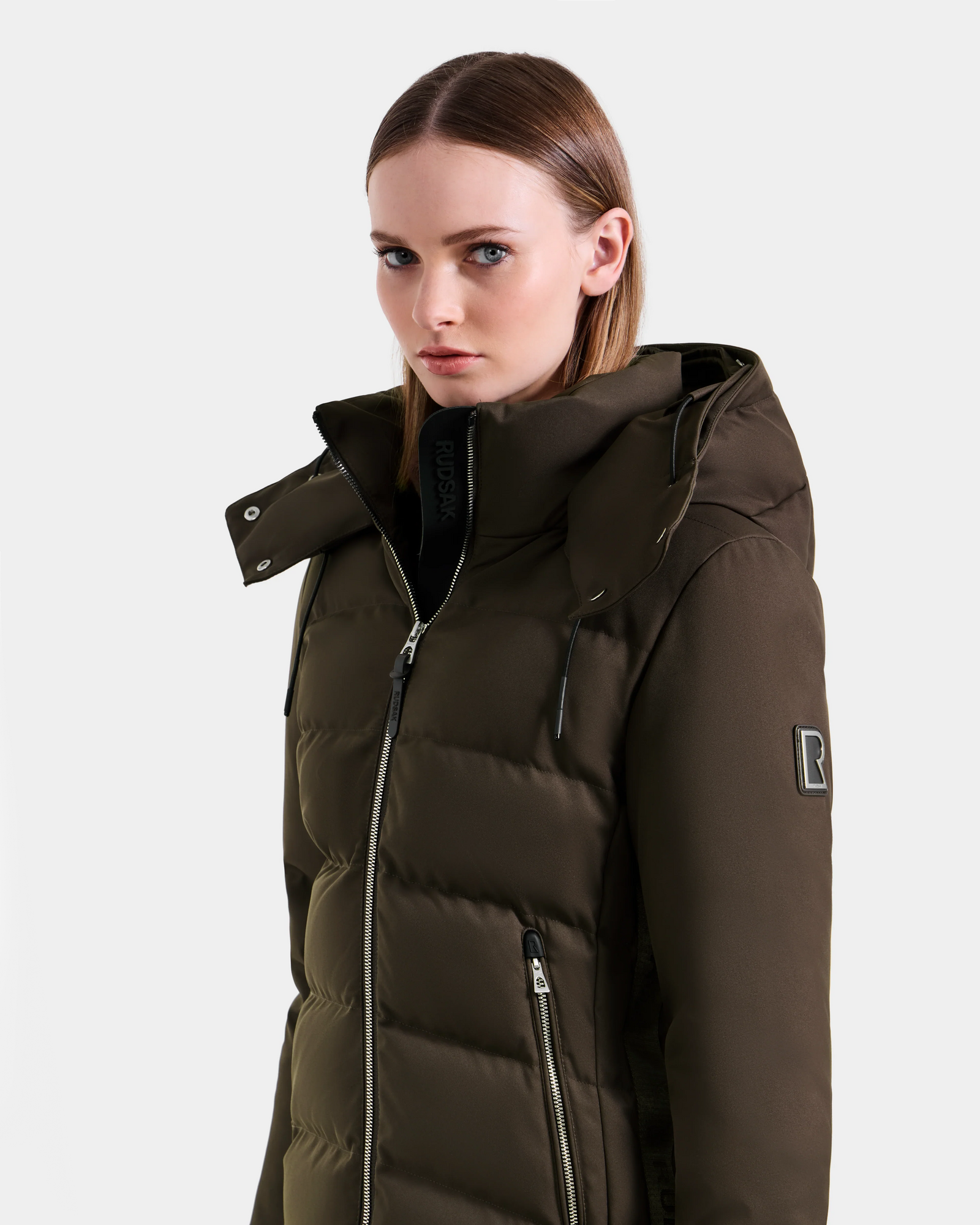 A woman with straight brown hair wears the Rudsak MIRRY M - L124533 Espresso women’s quilted down coat, featuring a hood, zippered front, and B patch on the sleeve, standing against a plain light background.