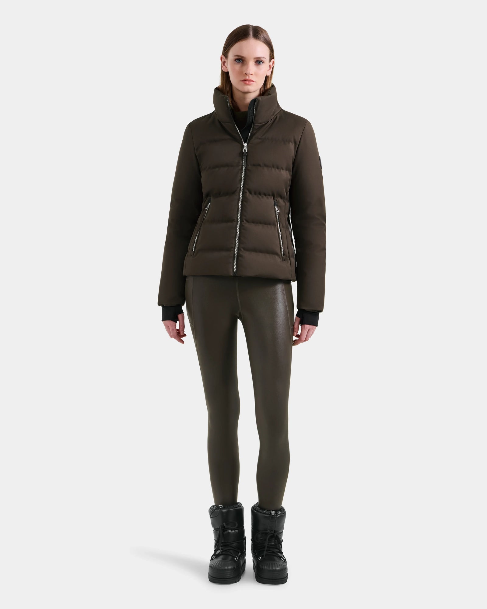 A woman stands facing forward in a Rudsak MIRRY M - L124533 Espresso RDS-certified down puffer, paired with fitted dark olive leggings and black winter boots against a plain white background.