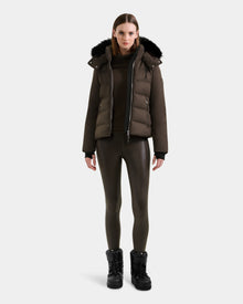 A woman faces forward in a Rudsak MIRRY M - L124533 Espresso quilted down coat with a black fur-trimmed hood, dark turtleneck, shiny brown leggings, and black boots against a plain light background.