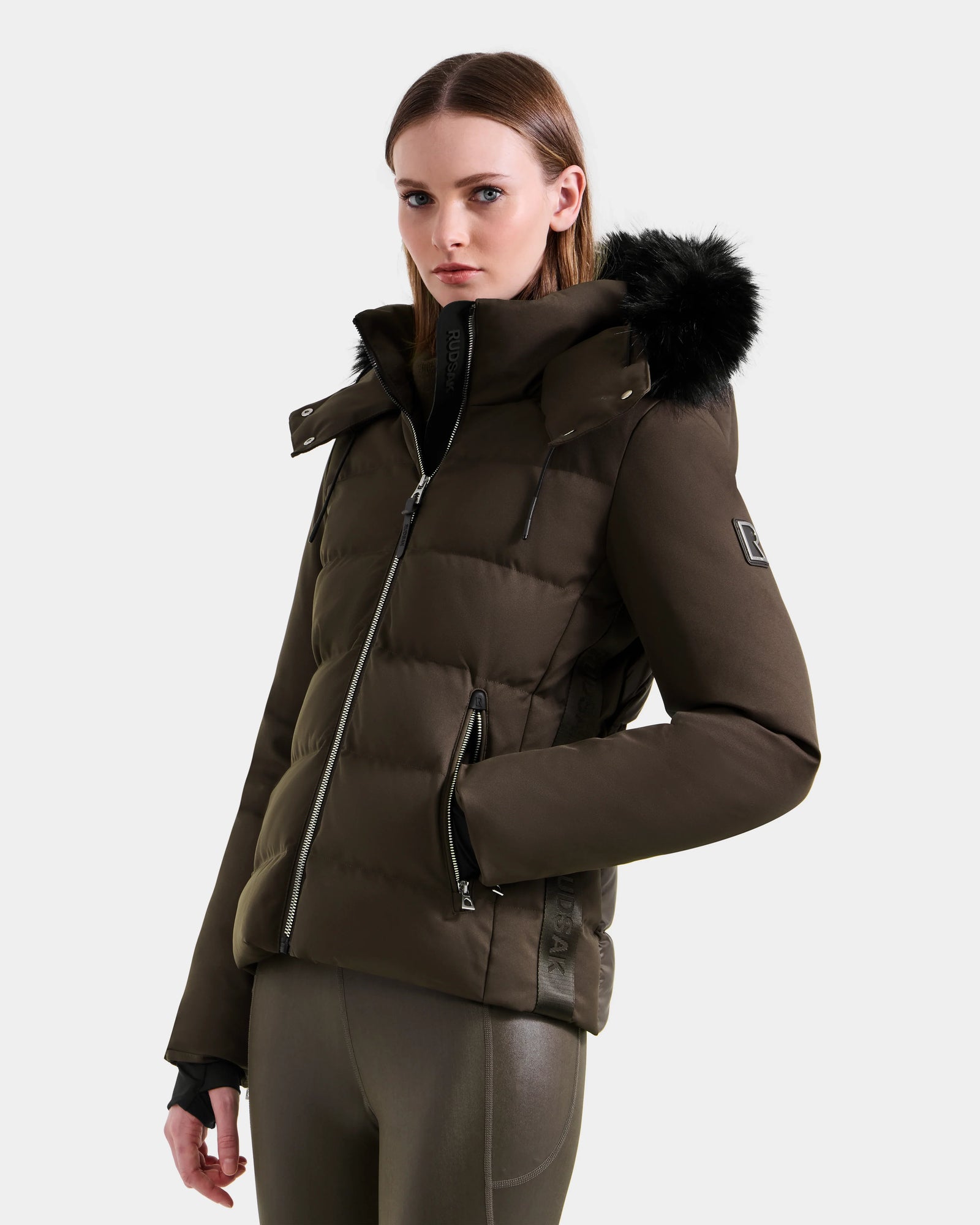 A woman stands against a plain light background, hands in pockets, wearing the Rudsak MIRRY M - L124533 Espresso women’s quilted down coat with a black fur-trimmed hood, looking toward the camera.